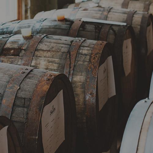 Rows of aged wooden whiskey barrels with white labels stacked in a distillery