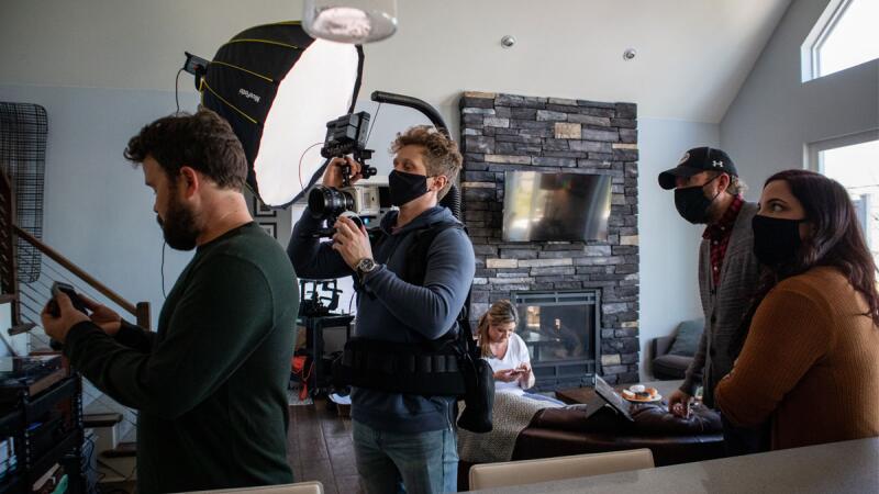 Masked film crew filming with a softbox light in a living room with a stone fireplace