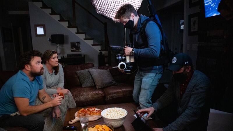 Masked videographer filming a couple watching TV with pizza and popcorn on the table