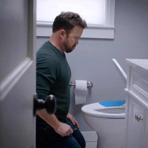 Man beginning to sit on a toilet fitted with a blue-accented Bemis seat