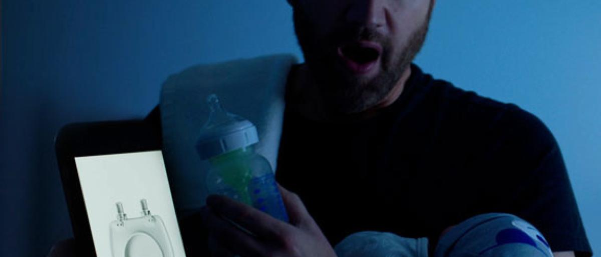 Man in a dark room looking at a phone showing a toilet seat while holding a baby and bottle