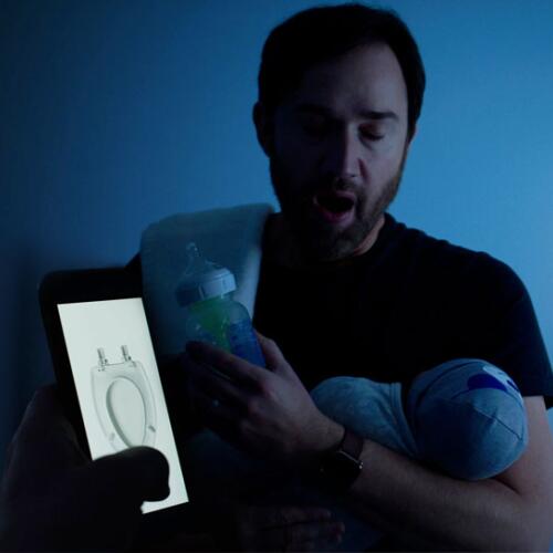 Man in a dark room looking at a phone showing a toilet seat while holding a baby and bottle
