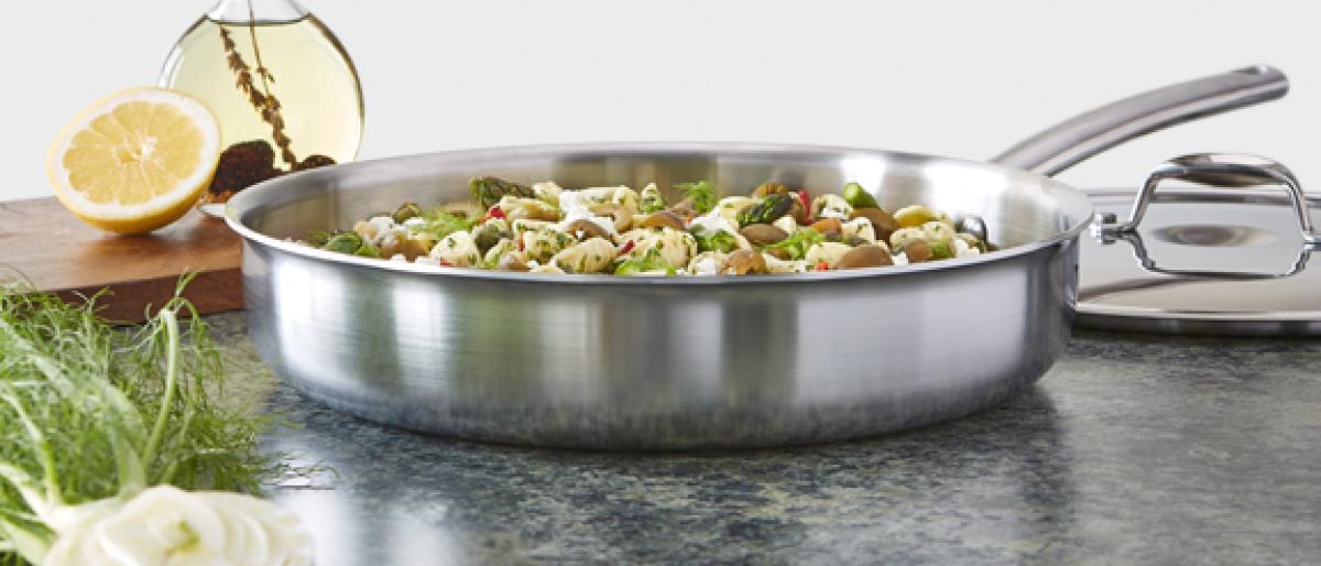 Stainless steel skillet with a pasta and vegetable dish, lemons, and olive oil on a counter