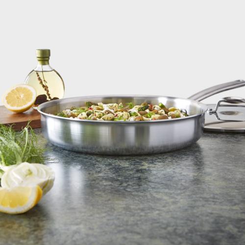 Stainless steel skillet with a pasta and vegetable dish, lemons, and olive oil on a counter