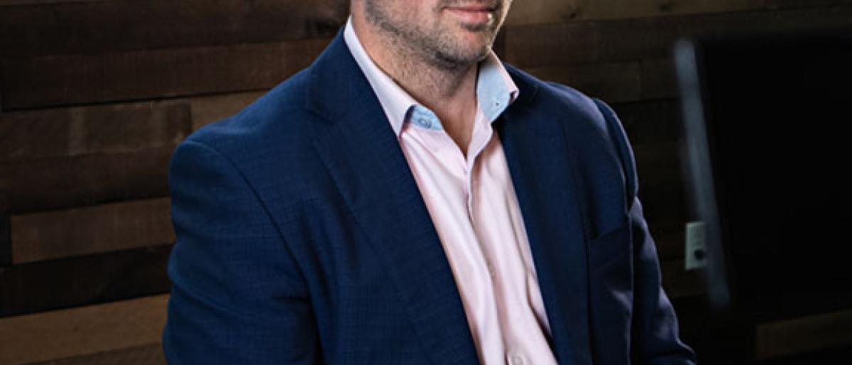 Man in a blazer and glasses seated for an on-camera interview against a wood-paneled wall