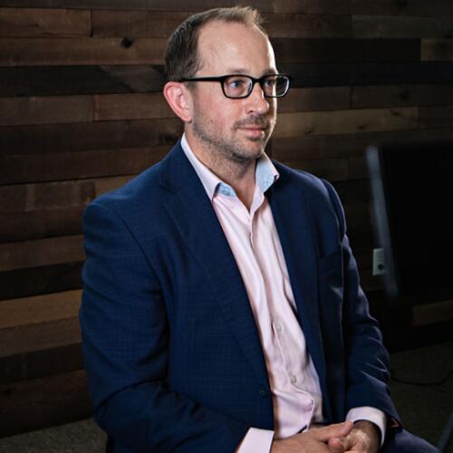 Man in a blazer and glasses seated for an on-camera interview against a wood-paneled wall